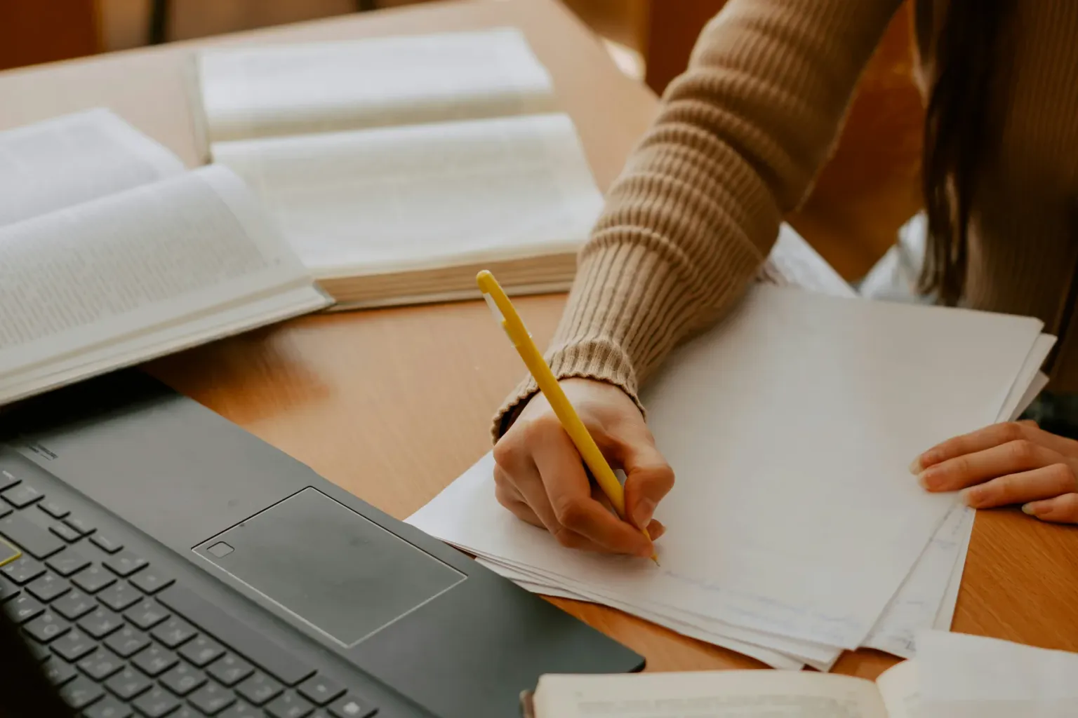 Person writing notes beside a laptop and open books
